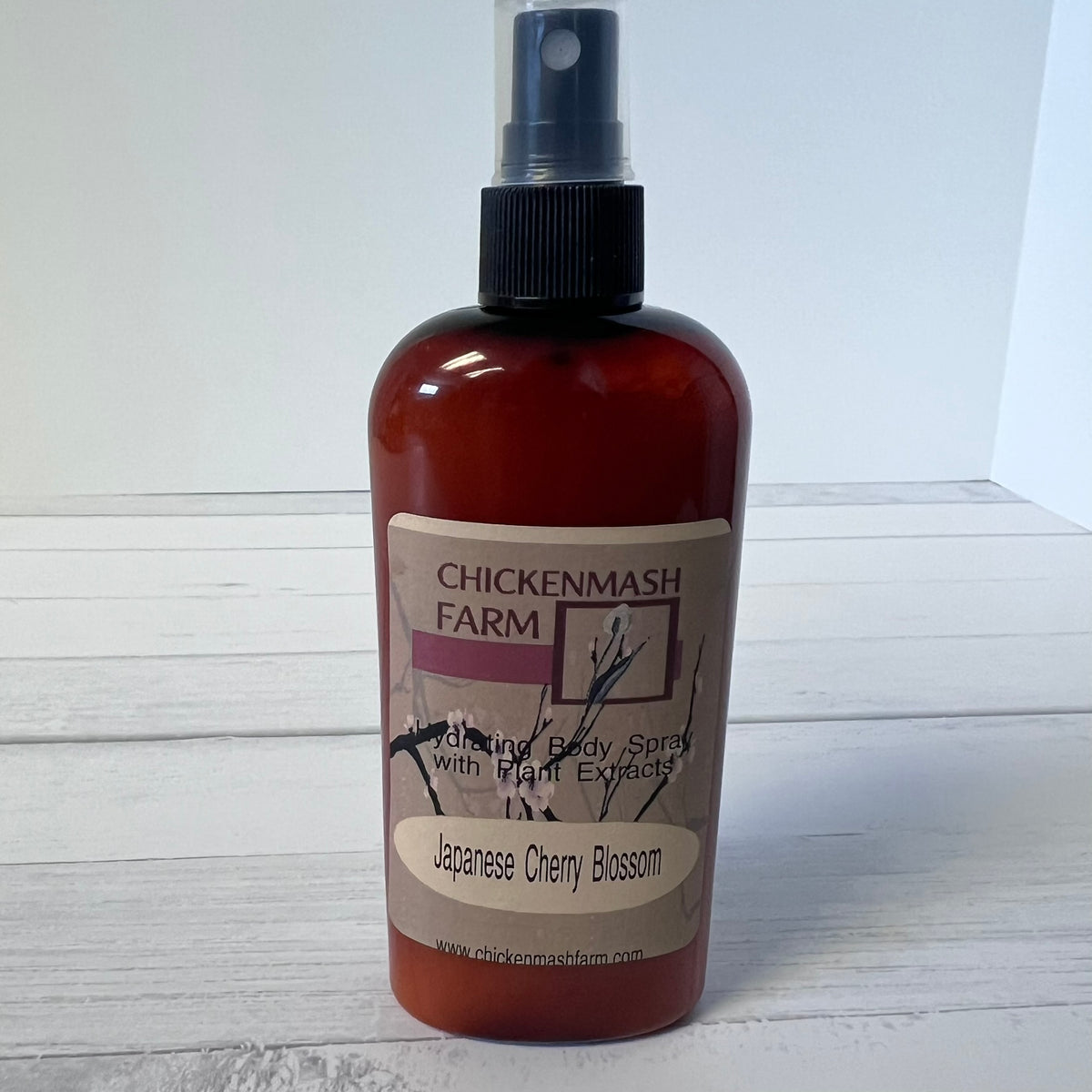 A bottle of Chickenmash Farm Japanese Cherry Blossom body spray with plant extracts, featuring a translucent amber-brown bottle and a black spray nozzle, set against a white wood plank background.