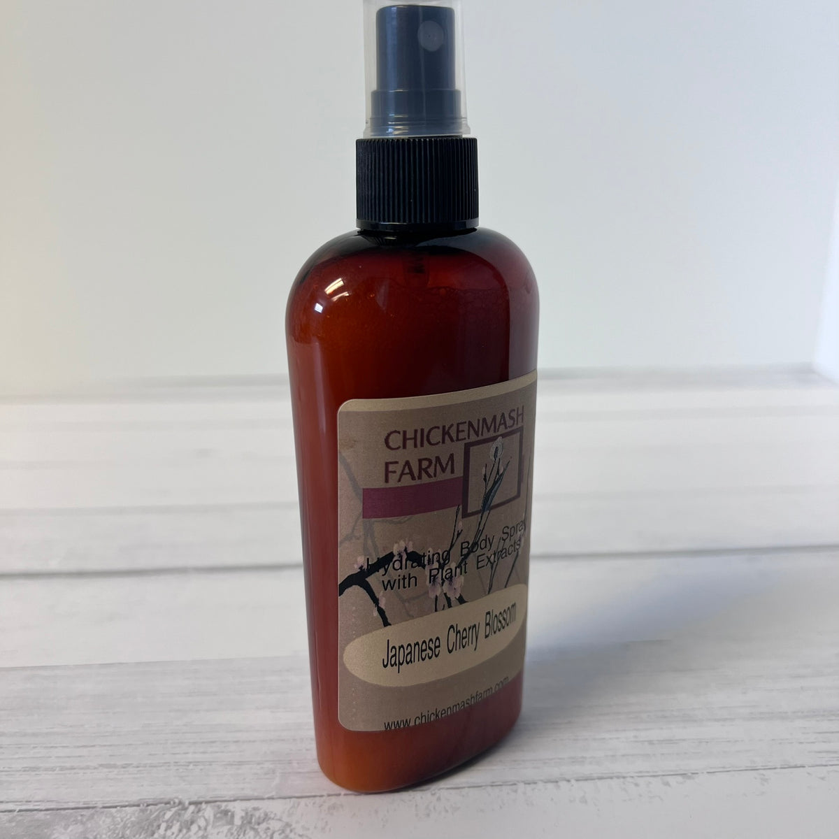 An amber plastic spray bottle with a black spray nozzle and a beige label reading 'CHICKENMASH FARM' and 'Japanese Cherry Blossom', standing on a white wooden surface against a white background.