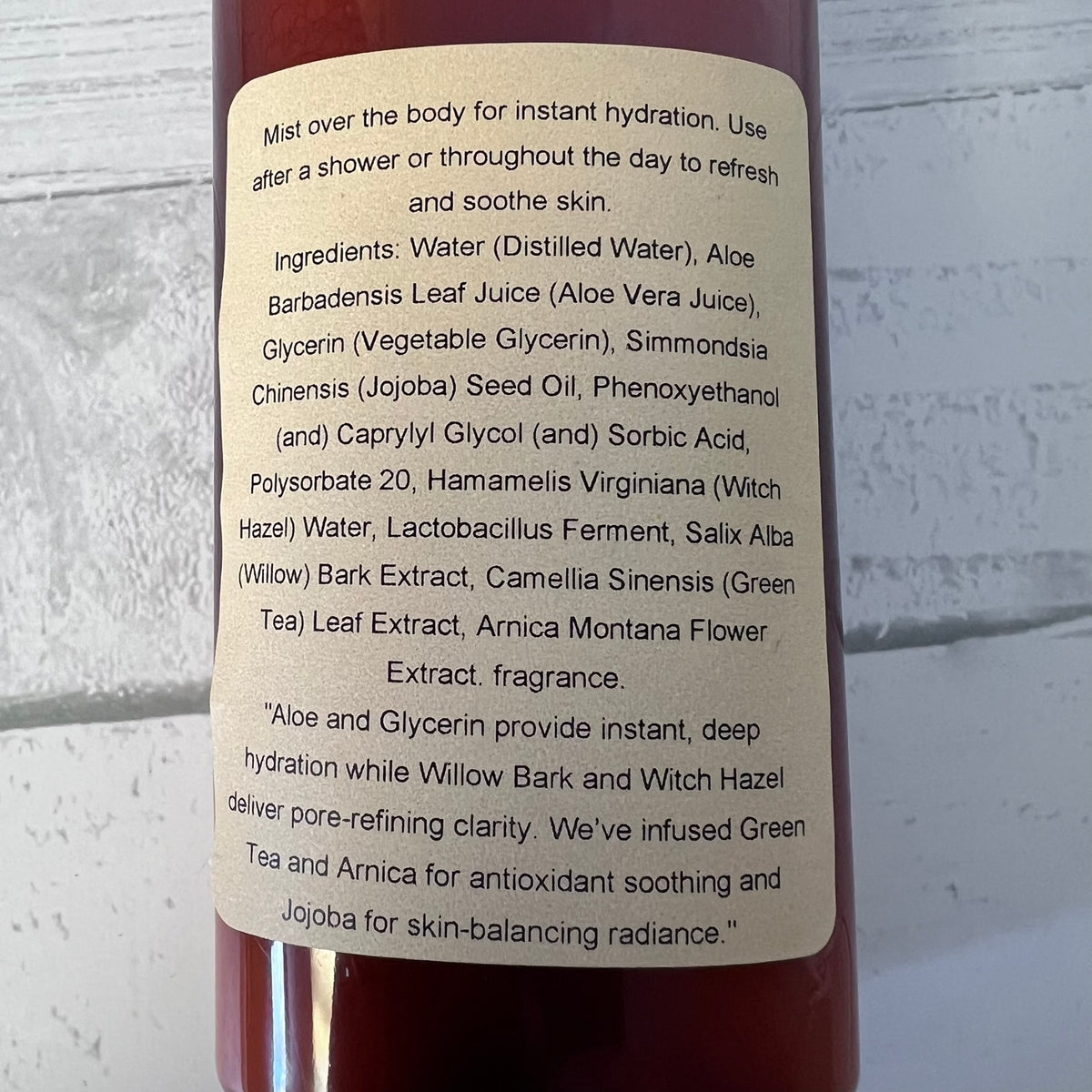 Close-up of a dark red bottle with a beige label showing product ingredients and usage text, photographed against a white painted brick wall.