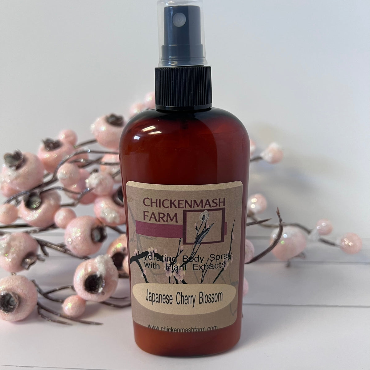 A bottle of Chickenmash Farm Body Spray with Plant Extract in Japanese Cherry Blossom scent, featuring an amber brown bottle and a beige label, styled with pink cherry blossom branches on a white surface.