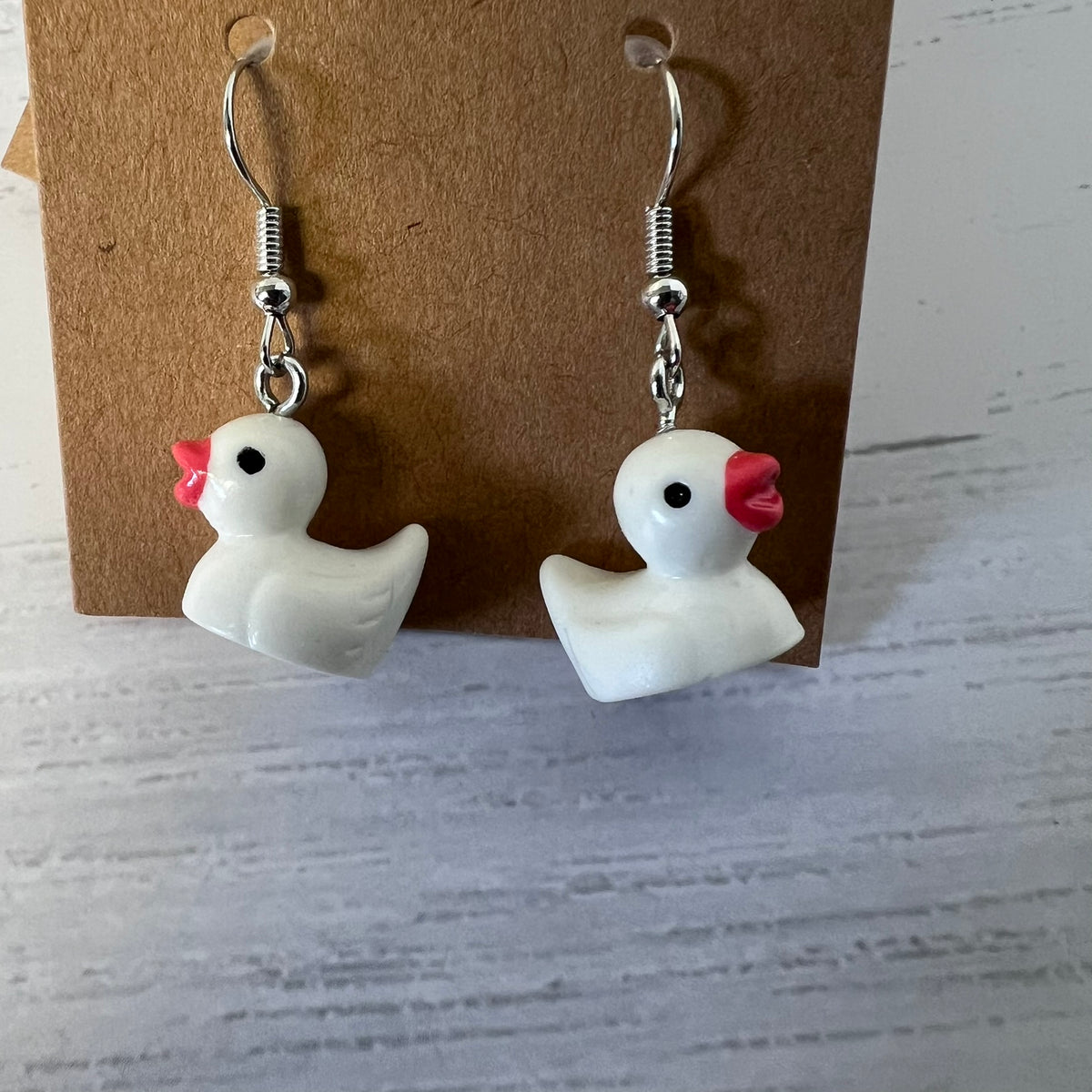 White duck-shaped earrings with red beaks on a brown card against a gray background