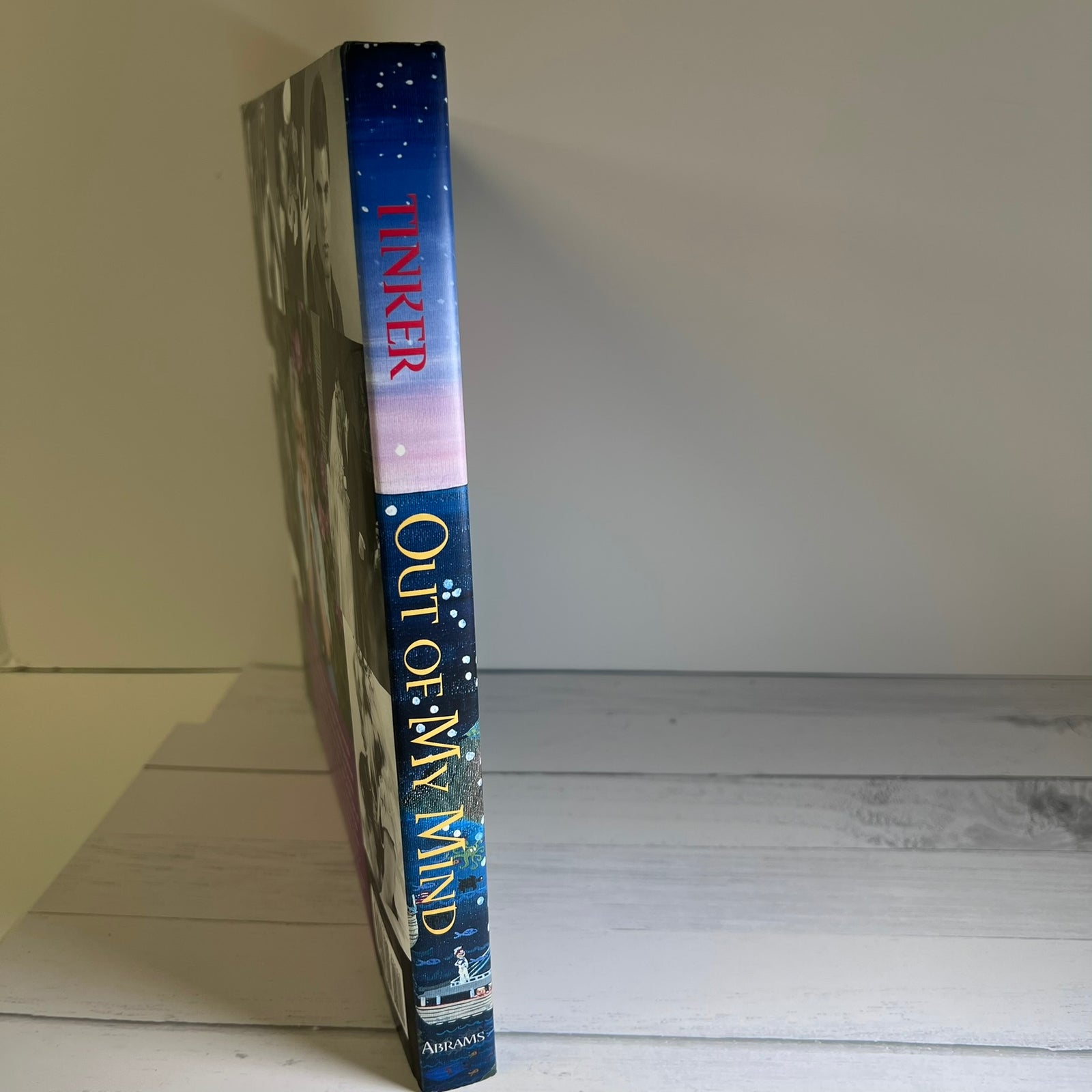 A hardcover book standing upright on a white wooden surface against a light gray wall. The spine is visible, showing the title 'OUT OF MY MIND', the author name 'TINKER' in red, and the publisher mark 'Amistad' at the bottom.