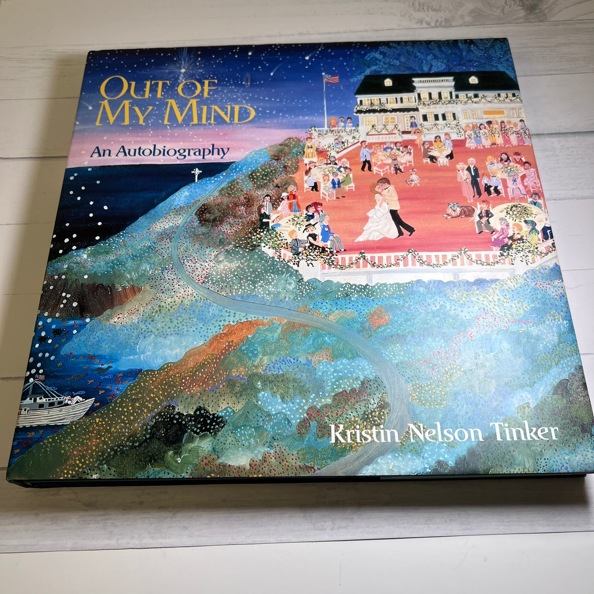 The book cover of 'Out of My Mind: An Autobiography' by Kristin Nelson Tinker, featuring a colorful, textured painting of a wedding scene on a hillside, photographed on a light wood surface.
