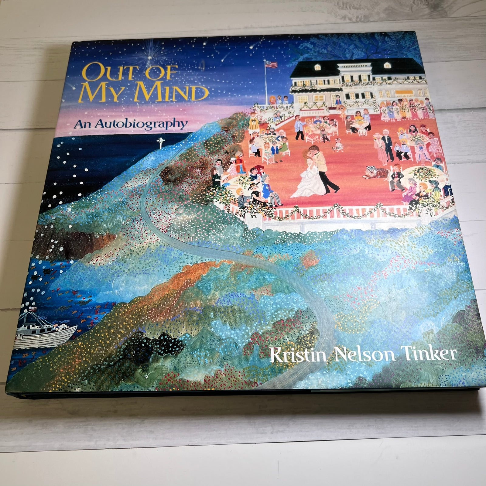 The book cover of 'Out of My Mind: An Autobiography' by Kristin Nelson Tinker, featuring a colorful, textured painting of a wedding scene on a hillside, photographed on a light wood surface.