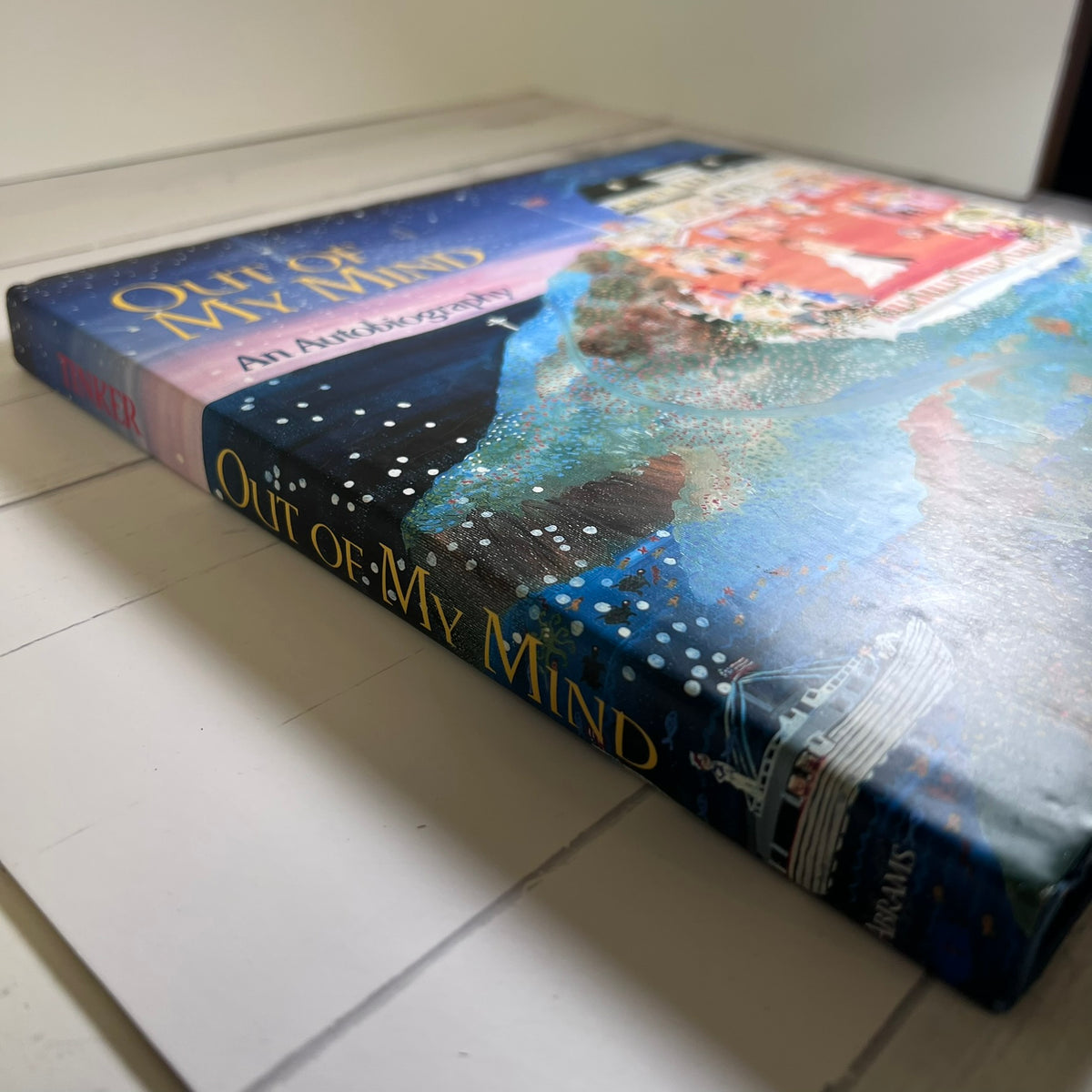 A hardcover book titled "Out of My Mind: An Autobiography" lying on a white surface. The cover features a colorful painted illustration with blue, purple, red, and white tones, and the title is printed in gold lettering on the spine and front cover.