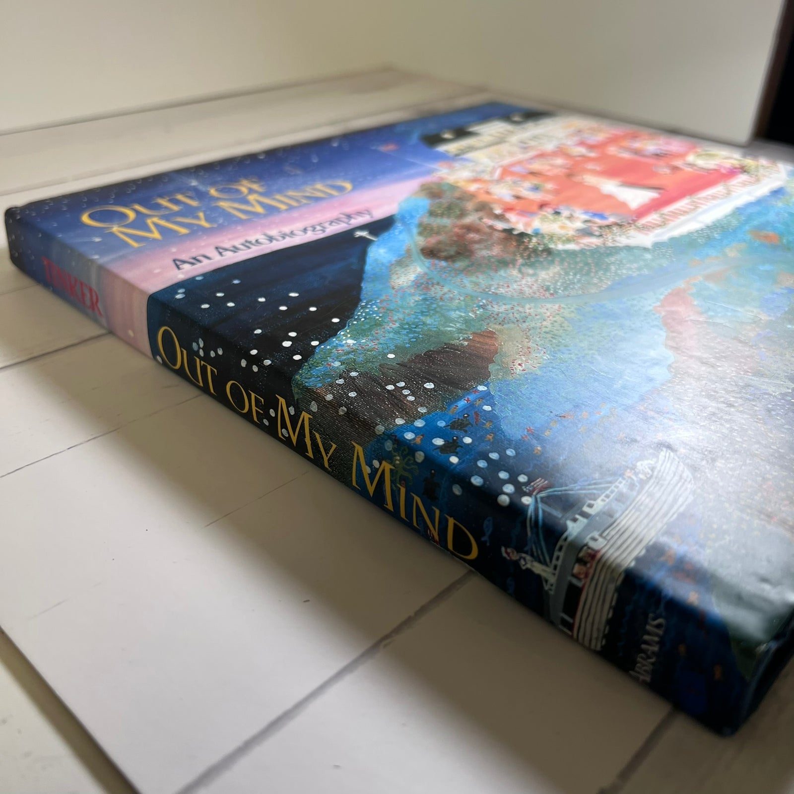 A hardcover book titled "Out of My Mind: An Autobiography" lying on a white surface. The cover features a colorful painted illustration with blue, purple, red, and white tones, and the title is printed in gold lettering on the spine and front cover.