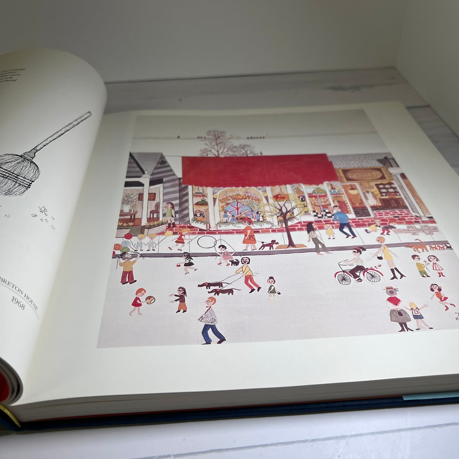 An open book showing a detailed vintage-style illustration of a bustling street scene with people, animals, bicycles, and storefronts. The page is photographed from above on a light surface.