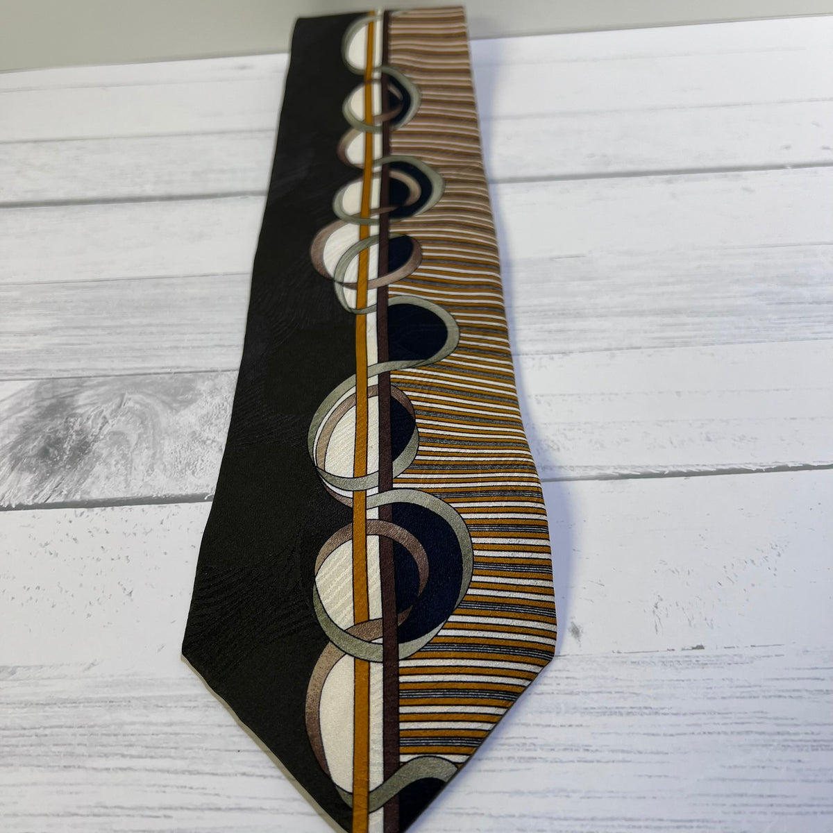 A patterned necktie laid flat on a white wood plank surface. The tie features a black left side with a vertical gold stripe and a row of overlapping circular motifs in black, white, and gold, transitioning into a right side with horizontal gold and white stripes.