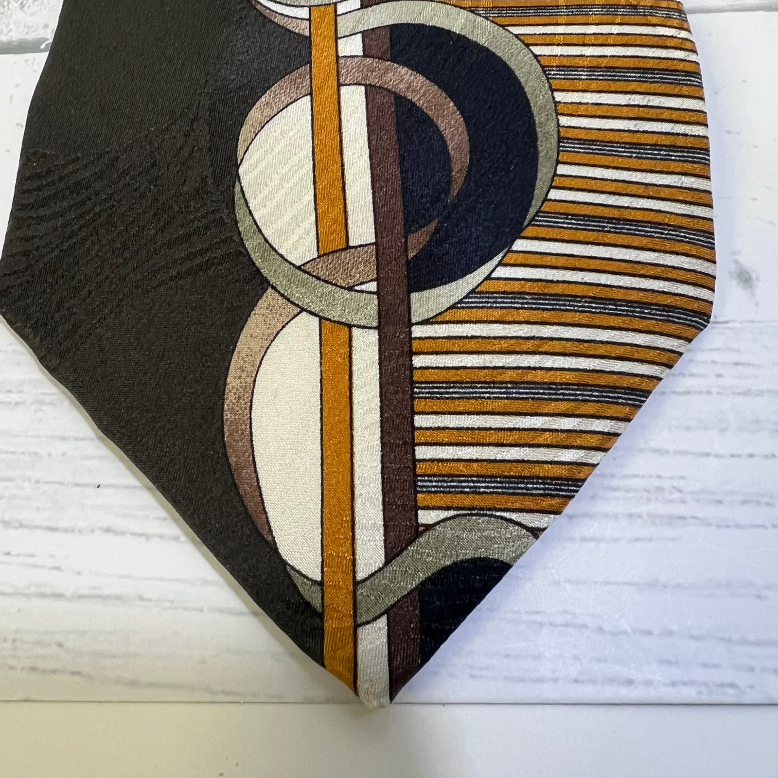 A close-up of a necktie featuring a modern abstract pattern with interlocking circles and stripes in black, brown, beige, white, and mustard yellow, laid flat on a white wood background.