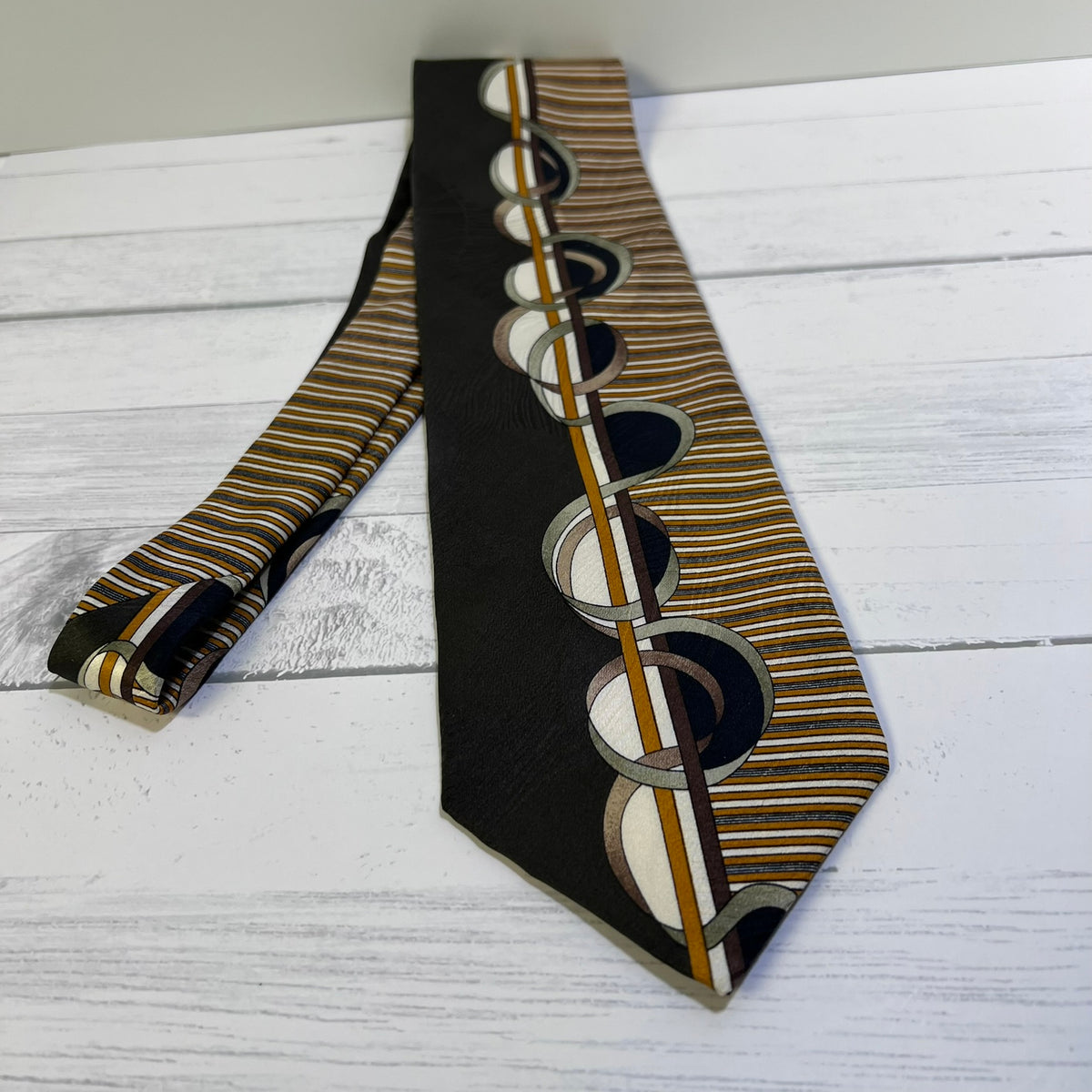 A patterned necktie laid flat on a white wood surface, featuring a black field with a vertical row of overlapping circular motifs in beige, brown, white, and black, alongside narrow tan and brown stripes.