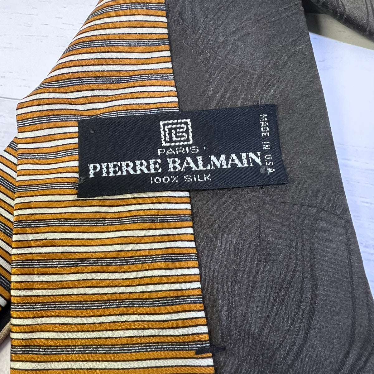 Close-up of a Pierre Balmain silk tie showing a striped pattern in mustard yellow, black, and white, alongside a dark gray smooth silk section and a black brand label.