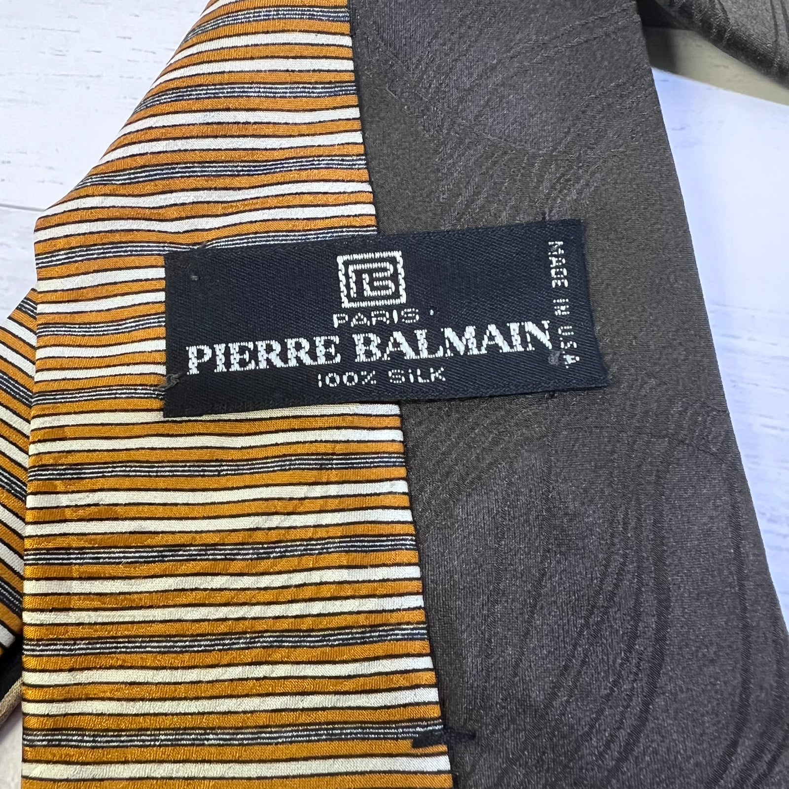 Close-up of a Pierre Balmain silk tie showing a striped pattern in mustard yellow, black, and white, alongside a dark gray smooth silk section and a black brand label.