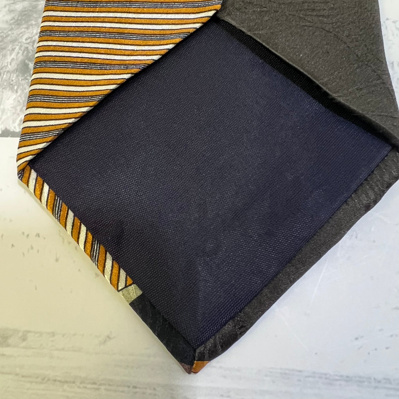 A folded necktie shown on a light surface, with one side displaying a navy blue satin front and the other side showing a striped pattern in mustard yellow, navy, and white.