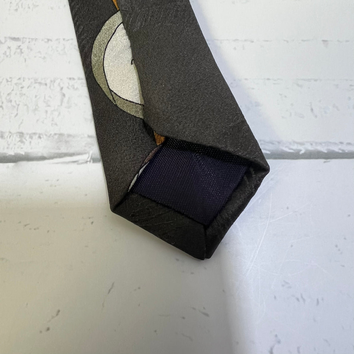 A close-up of a dark gray necktie with a circular moon pattern in beige and gray, folded to show a navy blue satin tip.