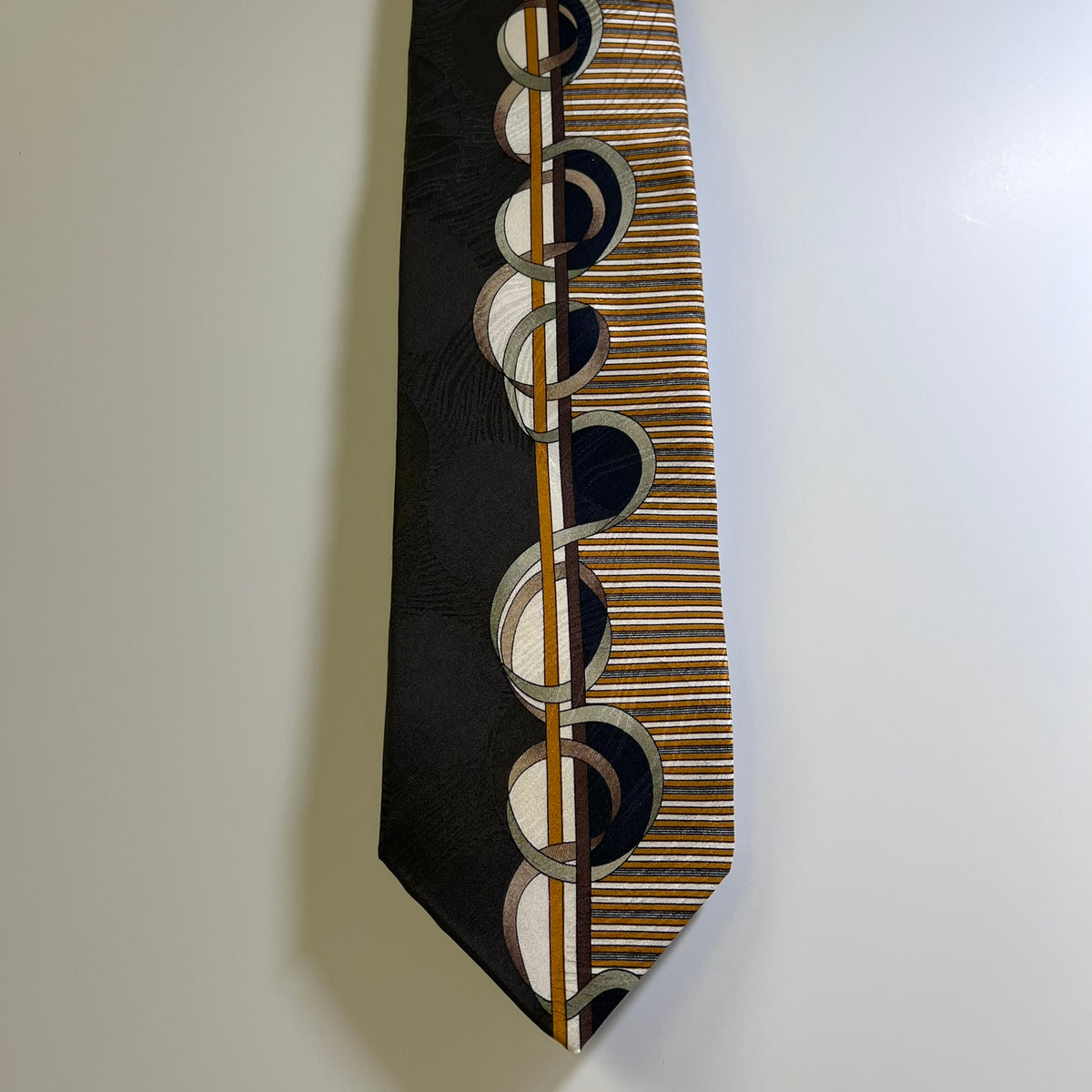 A men's necktie featuring a split design: one half is solid black with a subtle texture, and the other half has a pattern of gold, brown, and white stripes with overlapping circular motifs. The tie is displayed against a plain white wall.