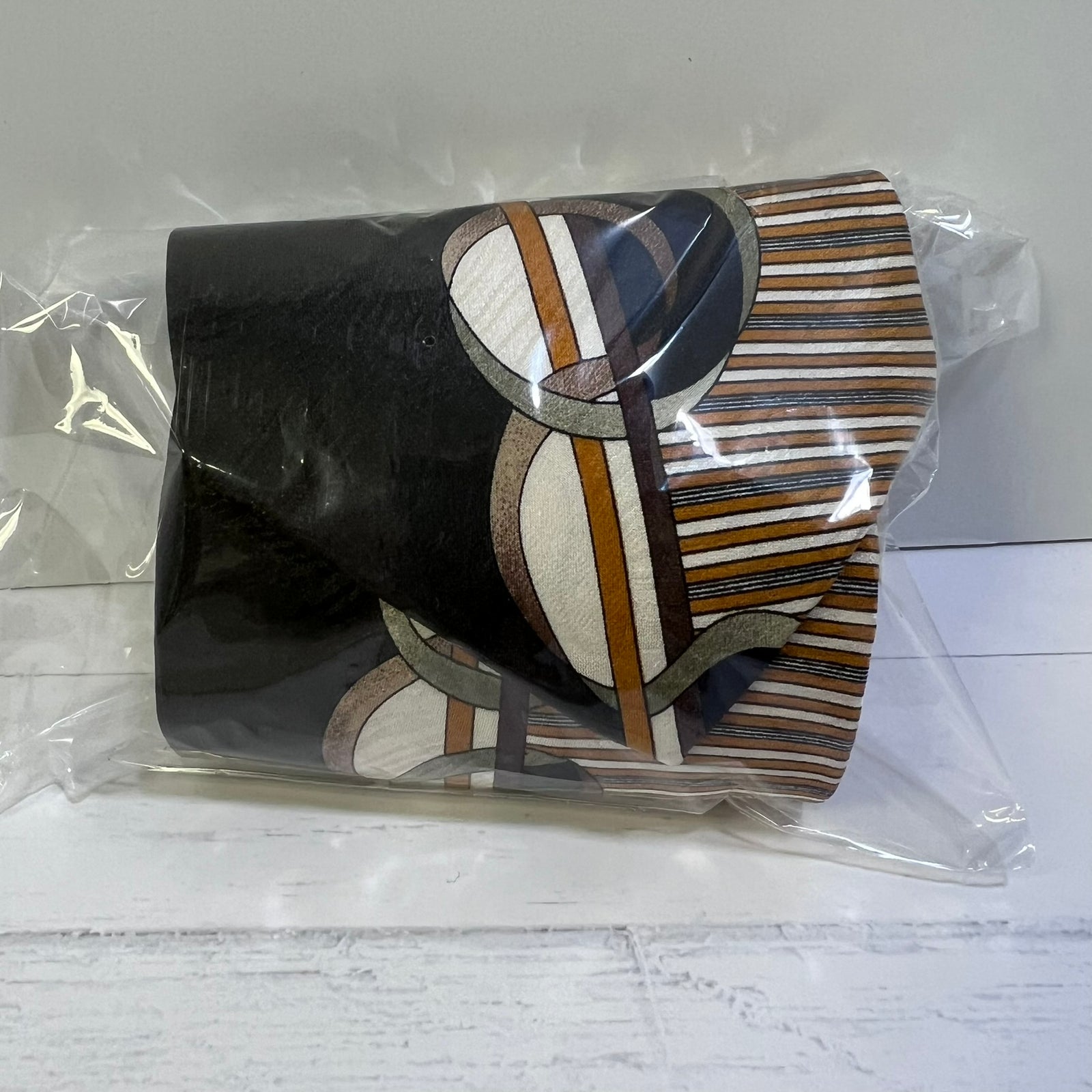 A rolled fabric or scarf-like product wrapped in clear plastic, featuring a black section and an abstract striped pattern in brown, beige, white, and gray.