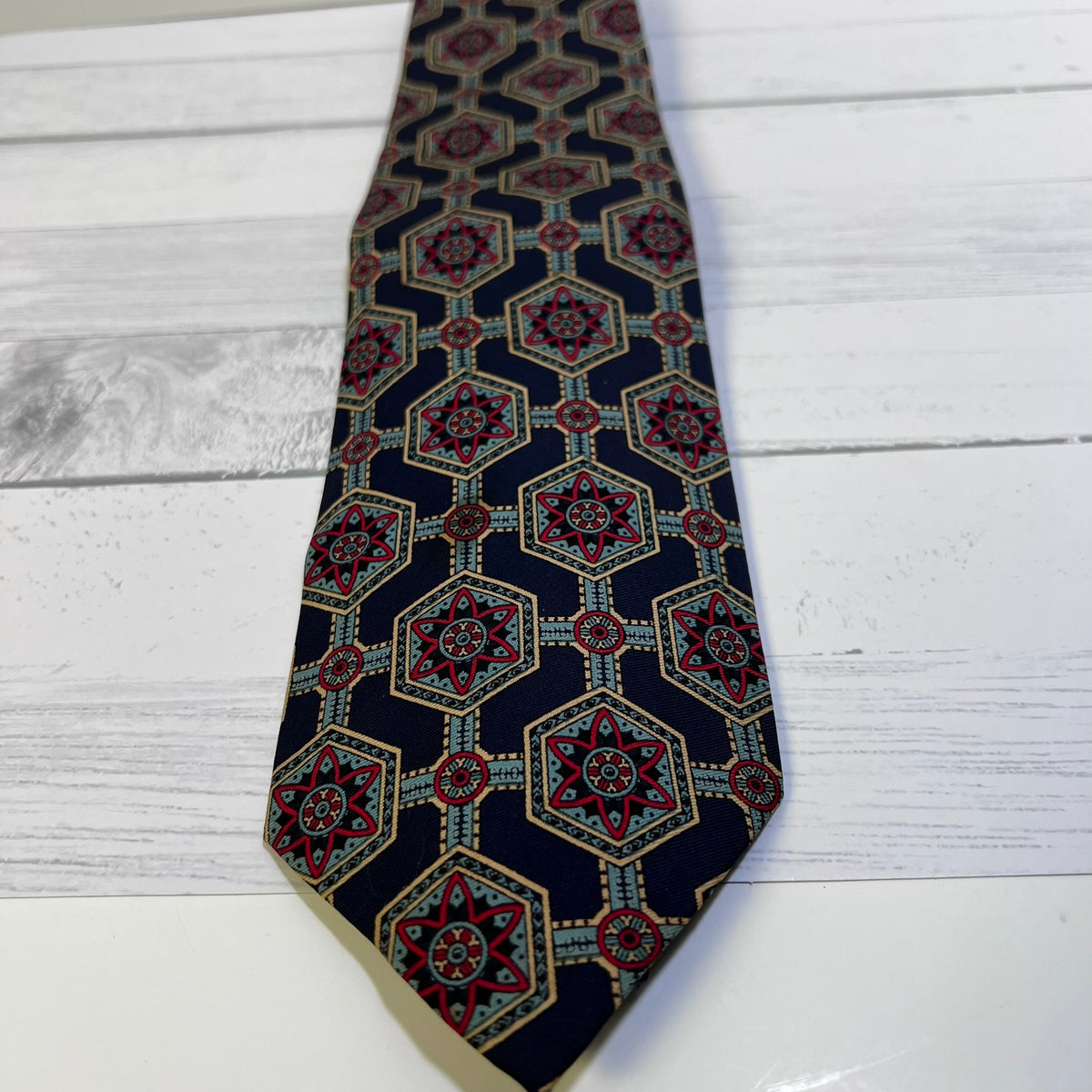 A patterned necktie with a navy blue base and a geometric medallion design in red, gold, and light blue, laid flat on a white wood plank surface.