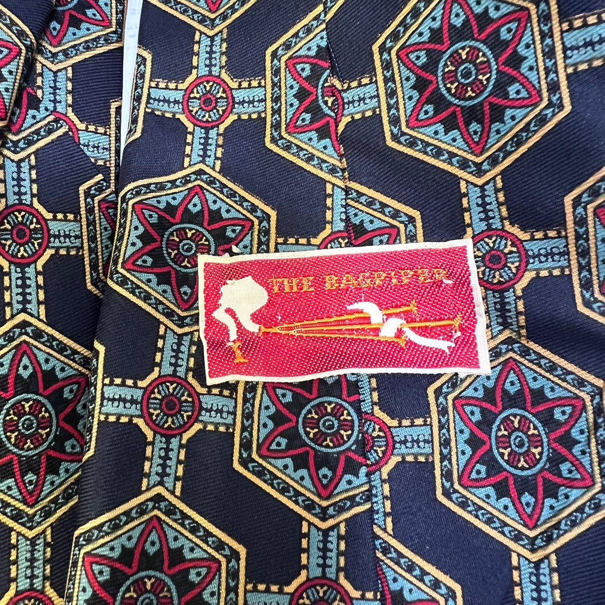 A close-up of a patterned necktie featuring a dark navy blue base with red, light blue, and gold geometric medallions. A red fabric brand label reading 'THE BARBER' is sewn onto the front.