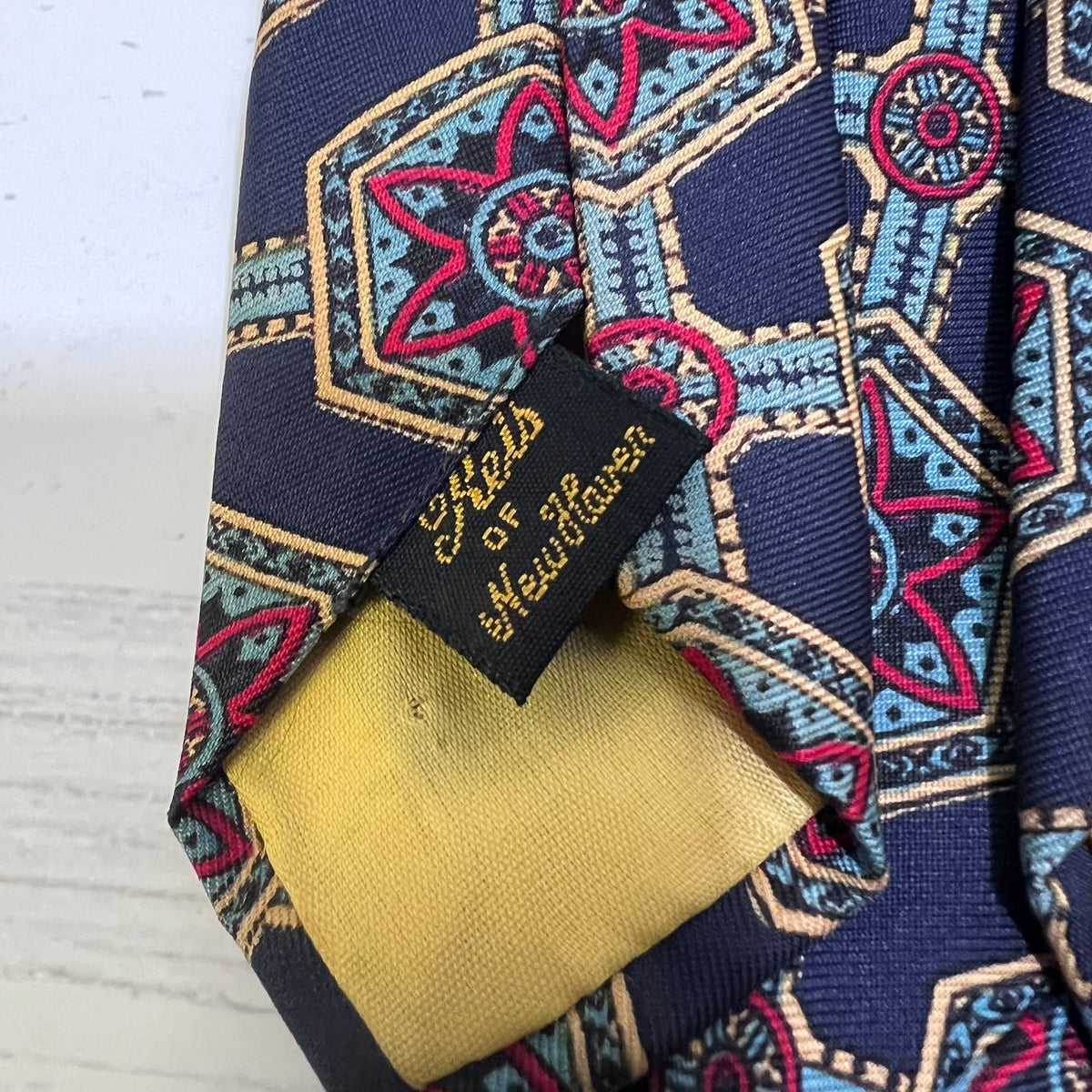 Close-up of a patterned necktie with a navy blue base, featuring geometric and star motifs in pink, light blue, and beige, with a black brand label reading 'Sheld of Heitorsten'.