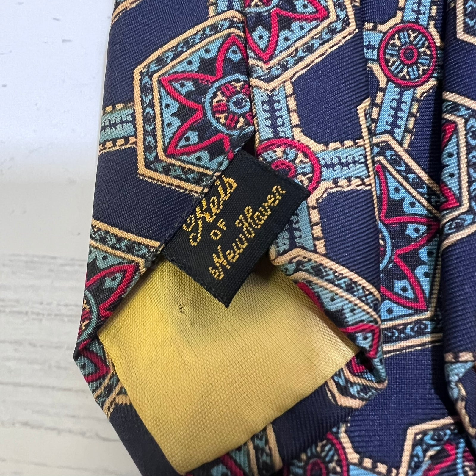 Close-up of a patterned necktie with a navy blue base, featuring geometric and star motifs in pink, light blue, and beige, with a black brand label reading 'Sheld of Heitorsten'.