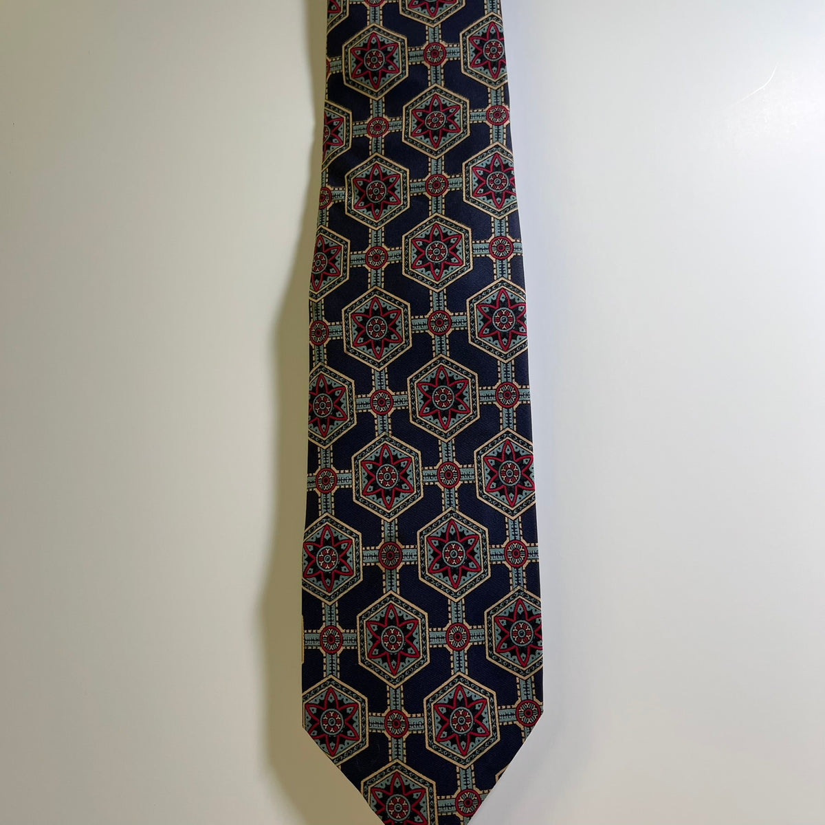 A patterned necktie with a dark navy blue base and a geometric medallion design in red, gold, and white, hanging against a plain off-white wall.