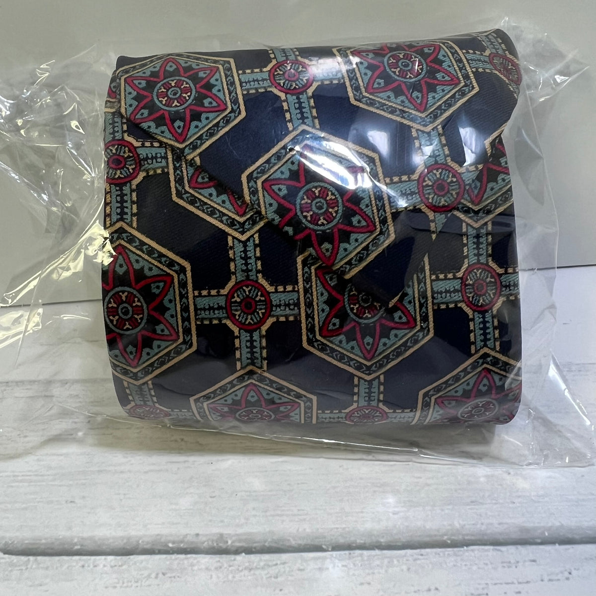 A rolled-up fabric accessory, likely a scarf or bandana, wrapped in clear plastic packaging. The fabric features a dark navy blue background with a geometric pattern in red, beige, and light blue.