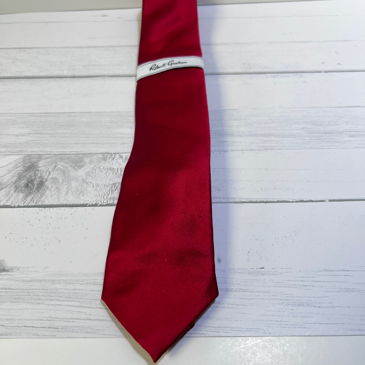 A red silk necktie with a white band inscribed with 'Robert Gordon' in black script, laid flat on a white wood plank background.