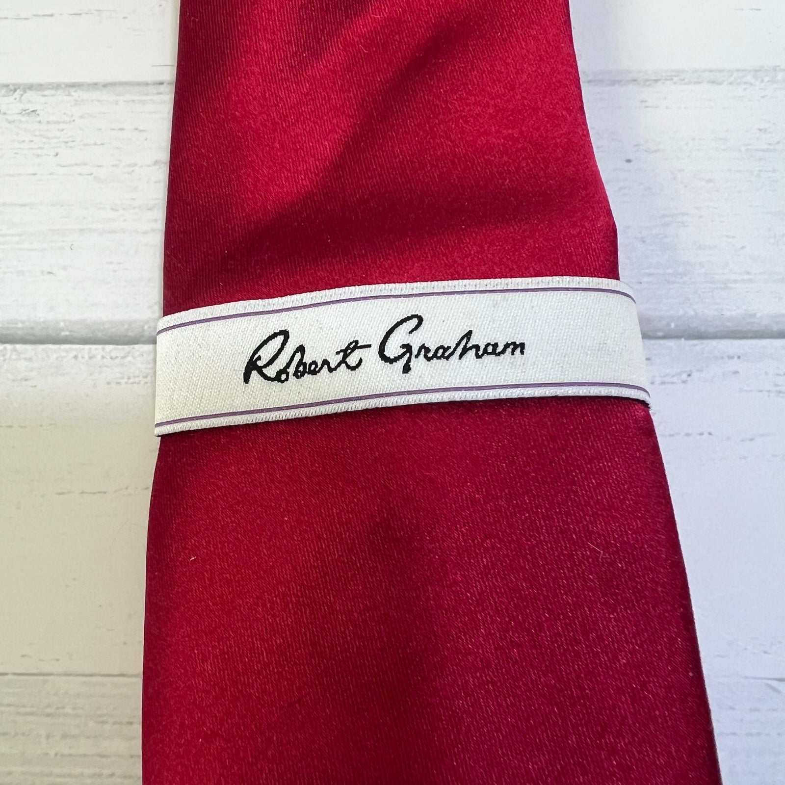 A close-up of a rolled red necktie with a white fabric band featuring the name 'Robert Graham' embroidered in black script, set against a white wood background.