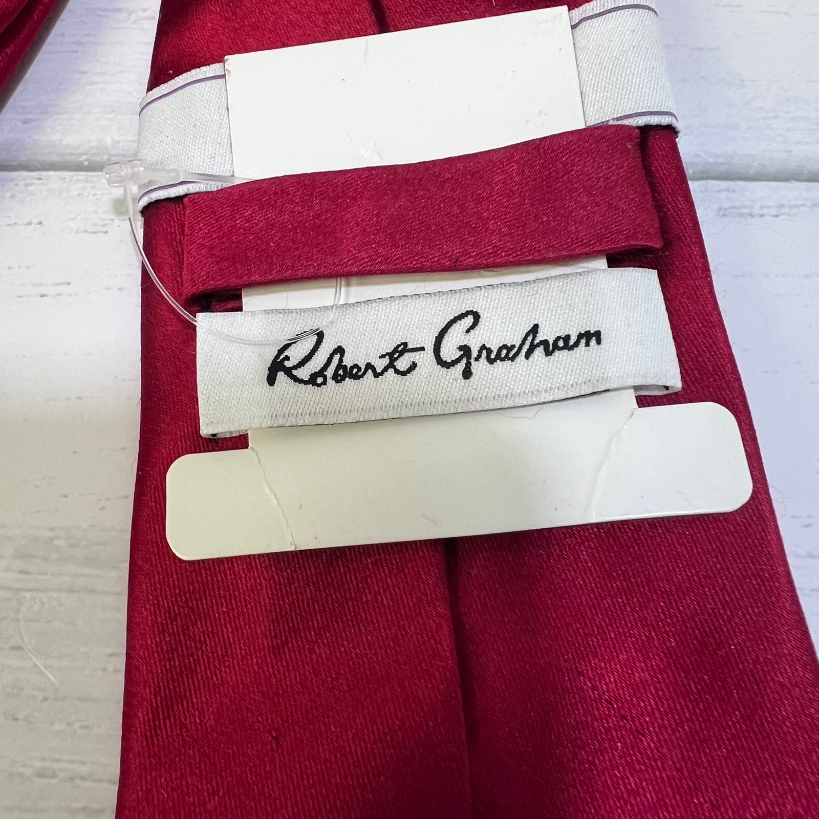 A close-up of a folded burgundy red necktie with white paper tags attached, featuring a white fabric brand label that reads 'Robert Graham' in black script.