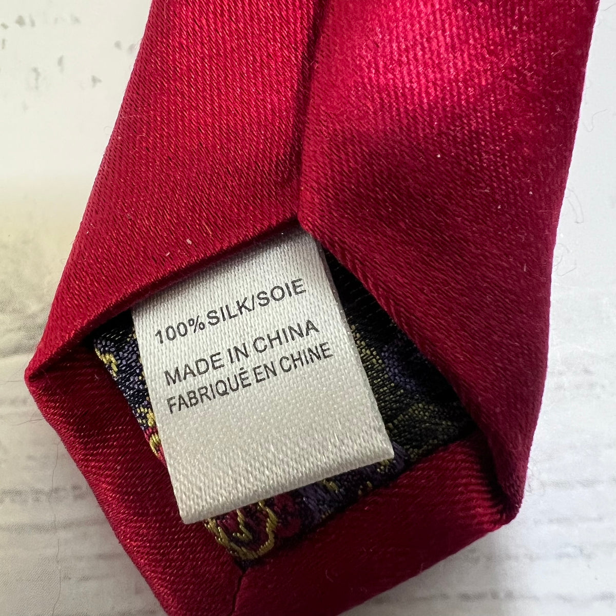 Close-up of a folded deep red silk necktie showing the inner patterned fabric and a white fabric tag that reads '100% SILK/SOIE MADE IN CHINA FABRIQUÉ EN CHINE'.
