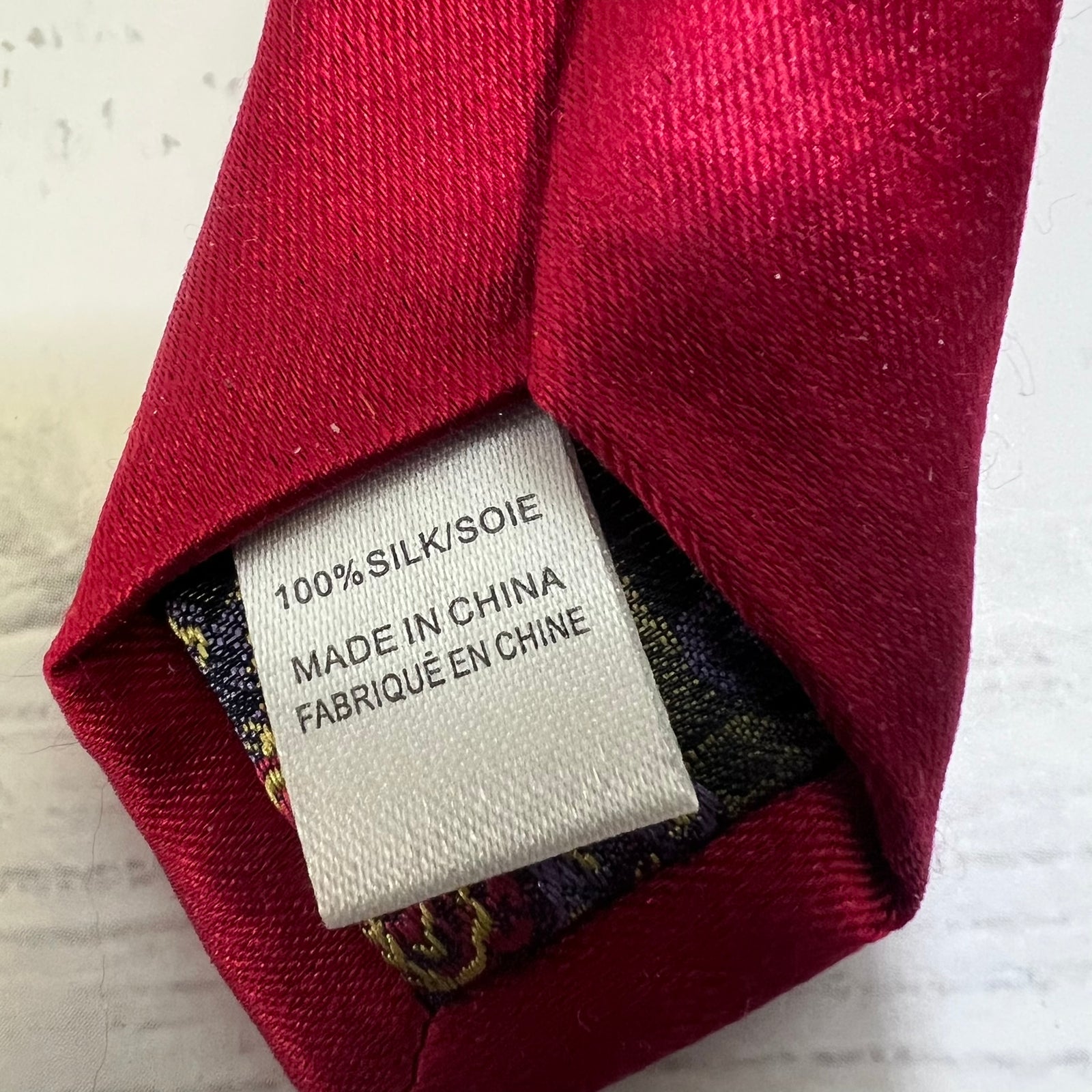 Close-up of a folded deep red silk necktie showing the inner patterned fabric and a white fabric tag that reads '100% SILK/SOIE MADE IN CHINA FABRIQUÉ EN CHINE'.