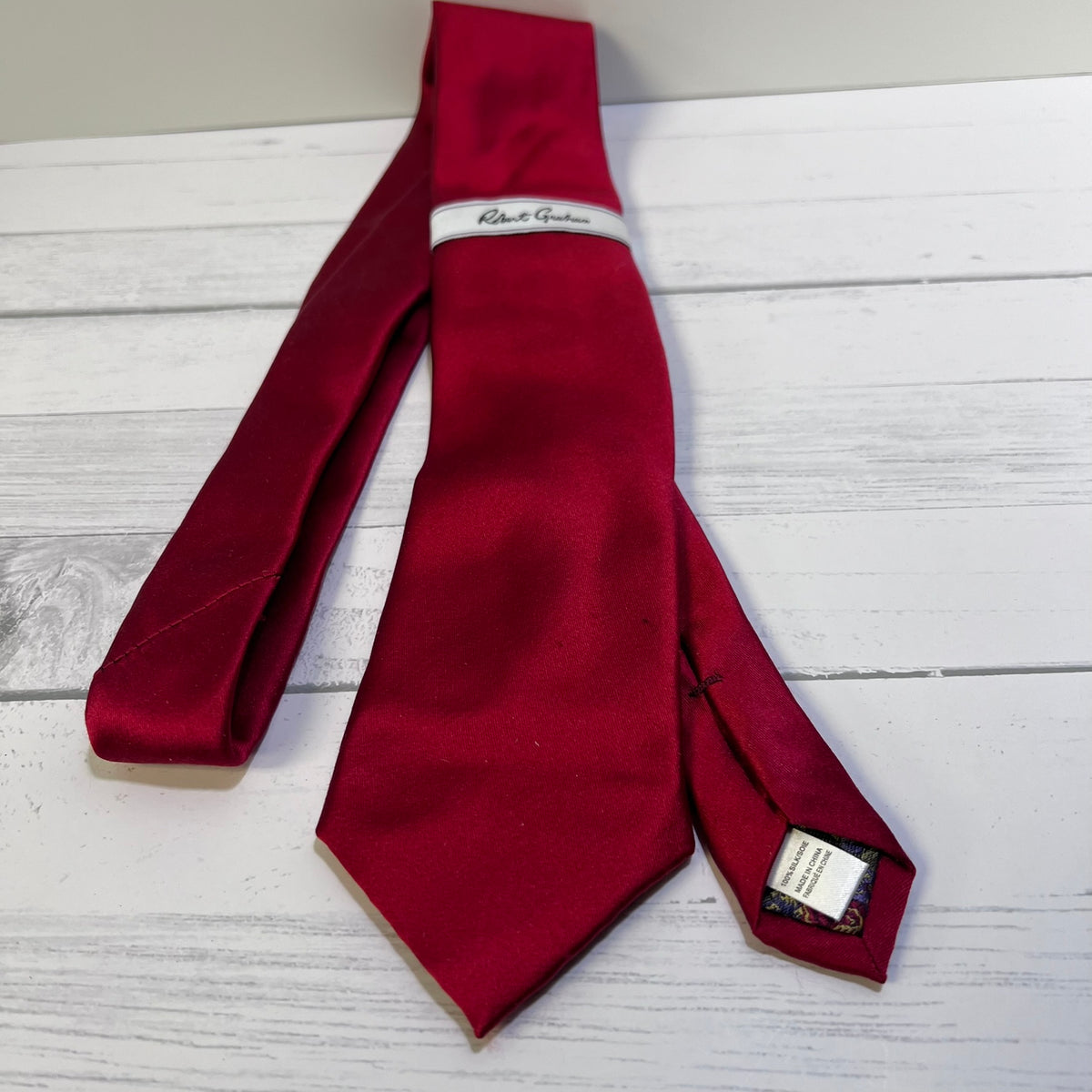 A deep red silk necktie laid flat on a white wood plank surface, with a white brand label reading 'Robert Graham' visible near the collar area.