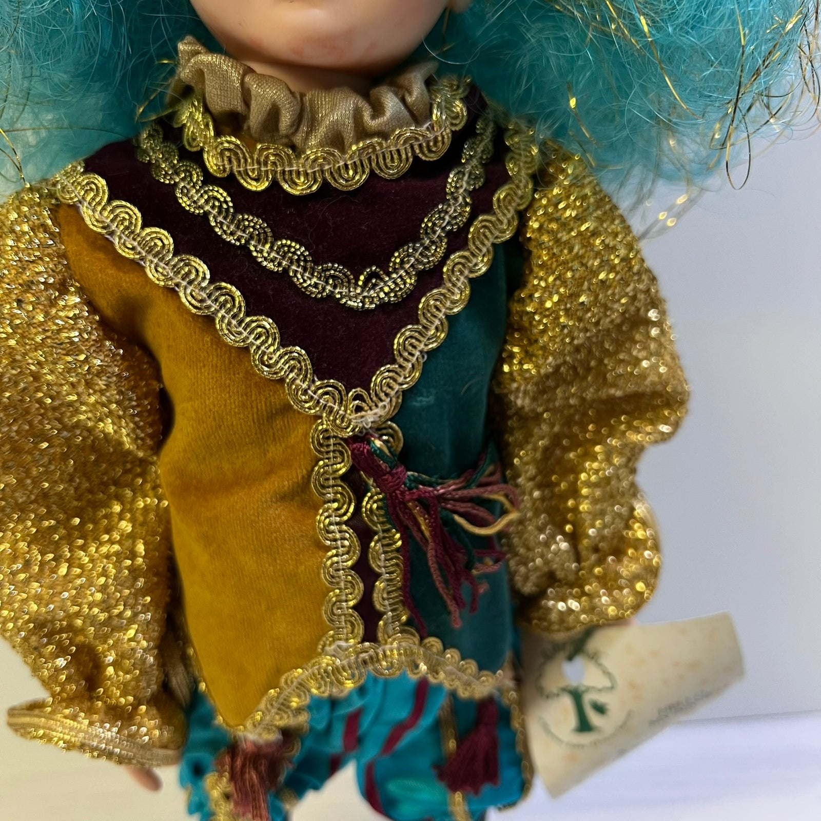 A close-up of a doll or figurine wearing an ornate costume with shiny gold ruffled trim, a mustard yellow bodice, teal and burgundy sections, and bright blue hair against a plain light background.