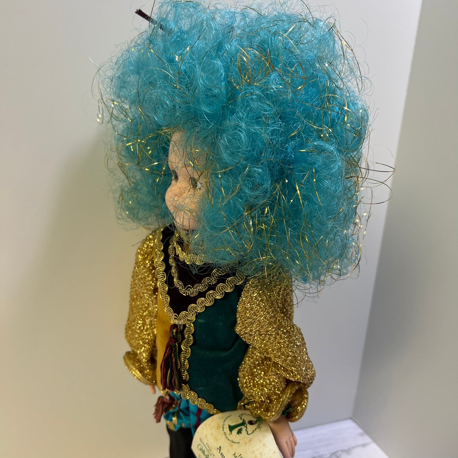 A collectible vintage doll with a large, voluminous curly turquoise blue wig with gold tinsel, wearing a glittering gold jacket over a dark green top. The doll is shown in profile against a plain light gray background.