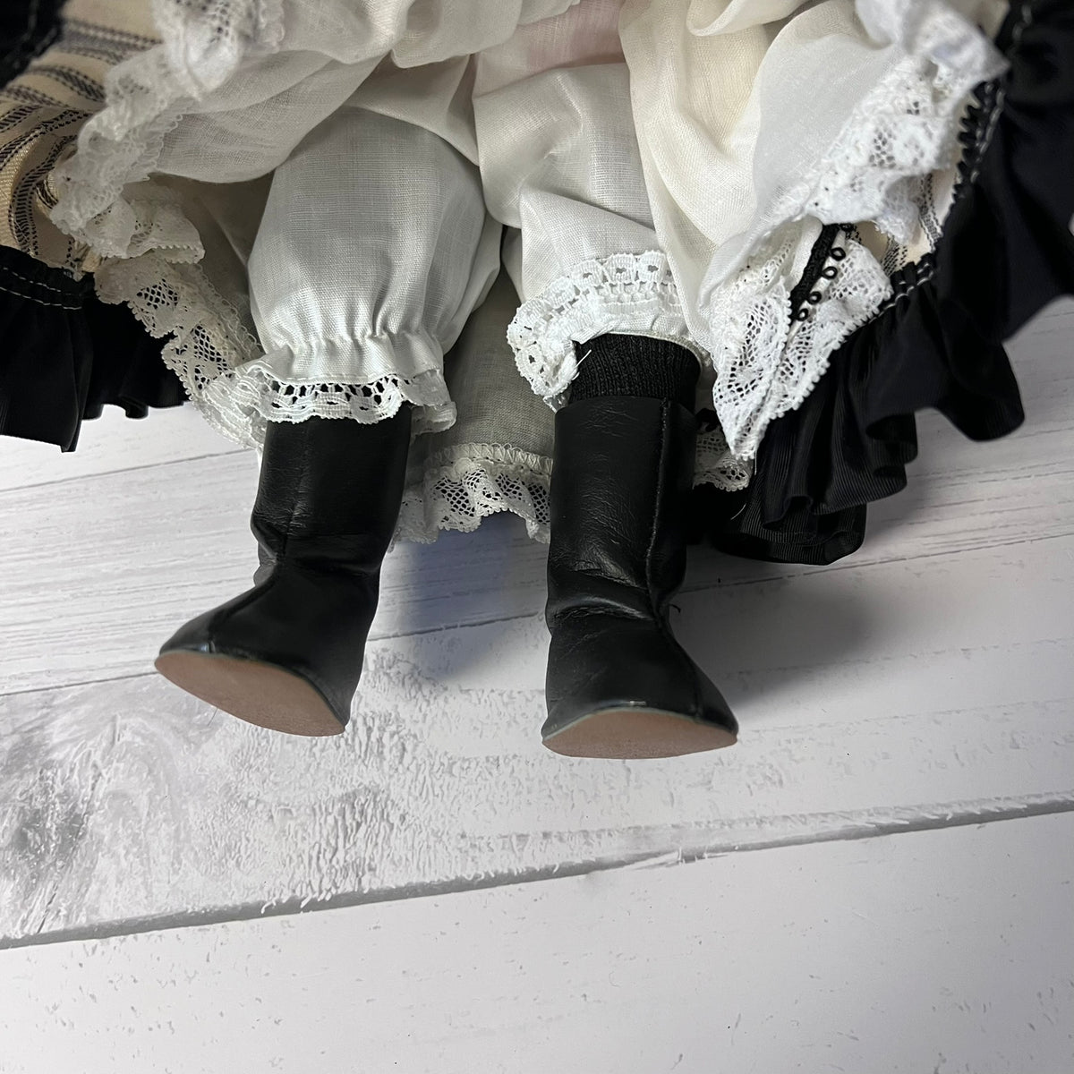Black leather doll boots with beige soles, shown on a white ruffled dress with lace trim, photographed from a top-down angle on a white wooden surface.