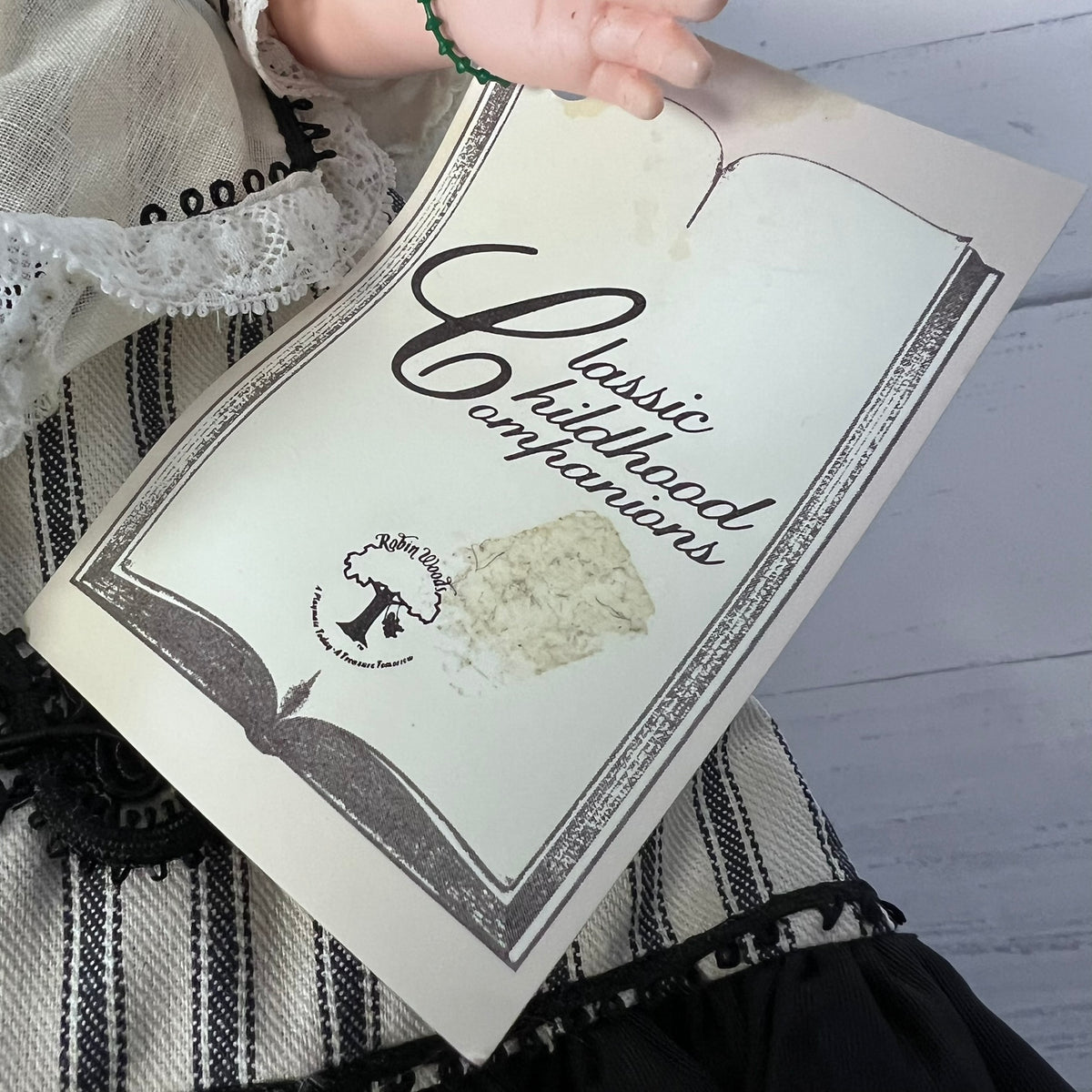 A hand holds an open vintage-style book or booklet titled "Classic Childhood Companions" over a black-and-white striped textile. The cover has an aged cream tone with dark script and border details, and a small circular logo near the lower left.