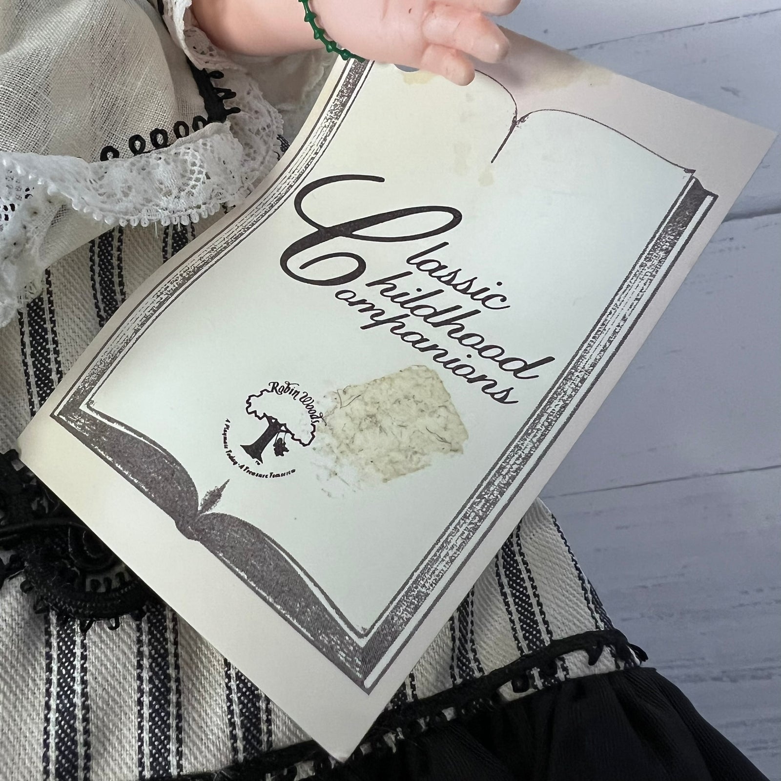 A hand holds an open vintage-style book or booklet titled "Classic Childhood Companions" over a black-and-white striped textile. The cover has an aged cream tone with dark script and border details, and a small circular logo near the lower left.