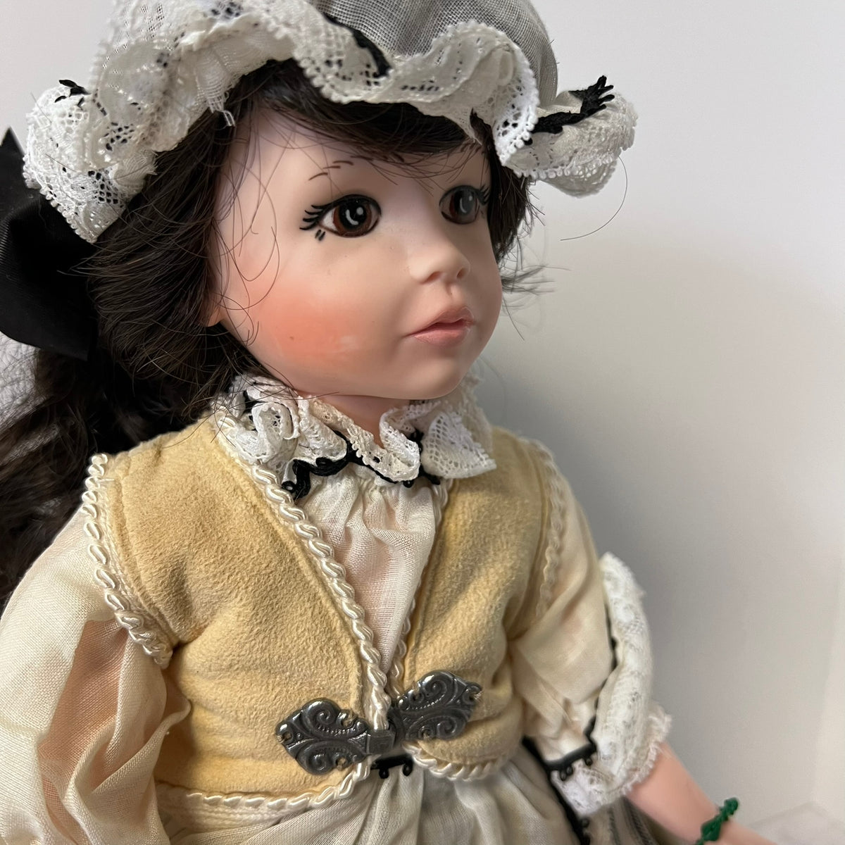 A close-up of a vintage-style porcelain doll wearing a cream and beige dress with lace trim, a gray bonnet, and dark brown hair, photographed against a plain light background.