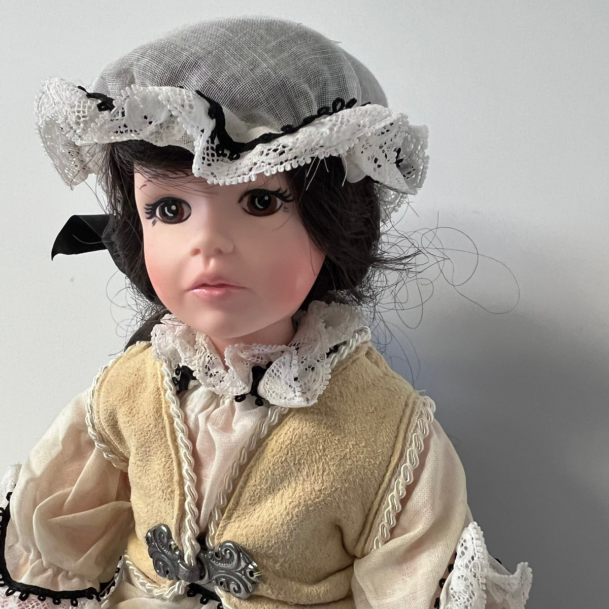 A doll with a porcelain face wearing a beige dress with white lace trim, a beige bonnet with white lace, and dark hair, photographed against a plain light gray background.