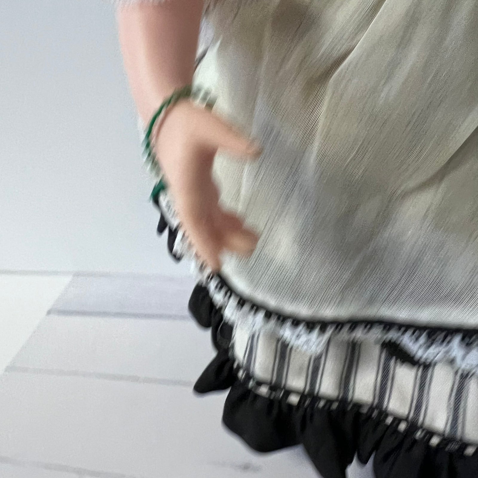 A close-up of a doll or mannequin torso wearing a shimmering champagne-silver top with a black and white striped ruffled hem and a green beaded bracelet on the wrist.