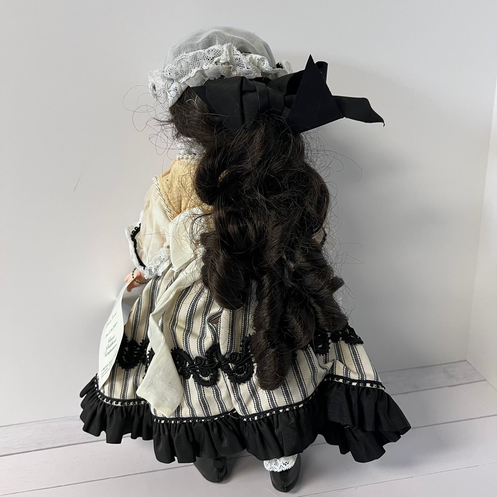 A vintage-style doll viewed from the back, wearing a cream and black striped dress with black lace trim, a black ribbon bow in its hair, and a gray bonnet.