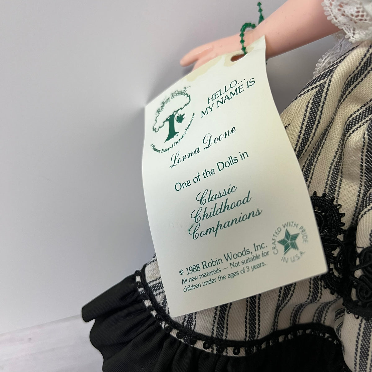 A close-up of a vintage doll tag attached to a striped black and beige dress, held by a doll's hand. The tag reads 'HELLO MY NAME IS Lorna Leone' and shows the Robin Woods logo.