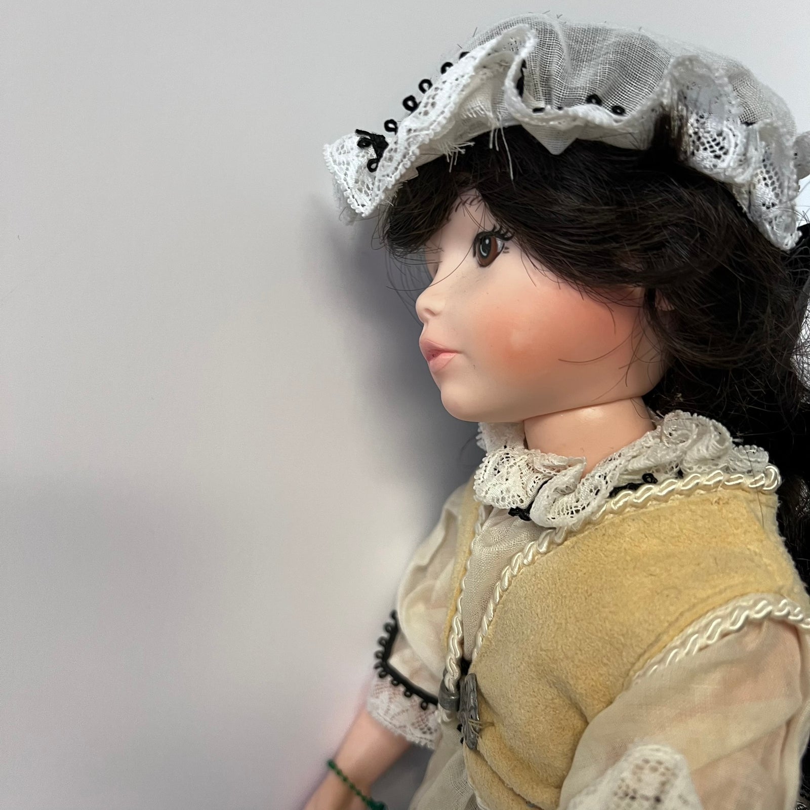 A vintage-style doll shown in profile against a plain light gray wall. The doll has dark brown hair, rosy cheeks, and is wearing a pale gray lace bonnet and a beige dress with white lace trim.