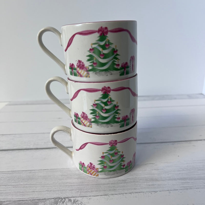 Stack of three Christmas-themed mugs with tree and gift designs on a white wooden surface.