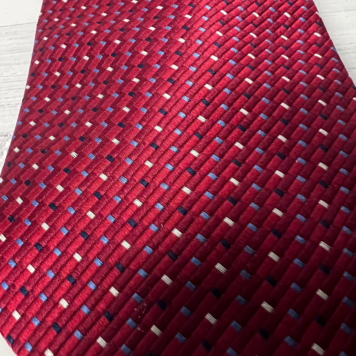 A close-up of a woven red necktie with a repeating pattern of small blue, white, and black accent threads.