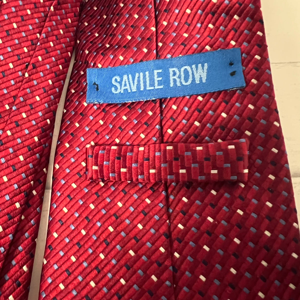 A close-up of a red textured necktie with a woven geometric pattern of blue, white, and black threads. A blue brand label reading 'SAVILE ROW' is visible near the collar area.