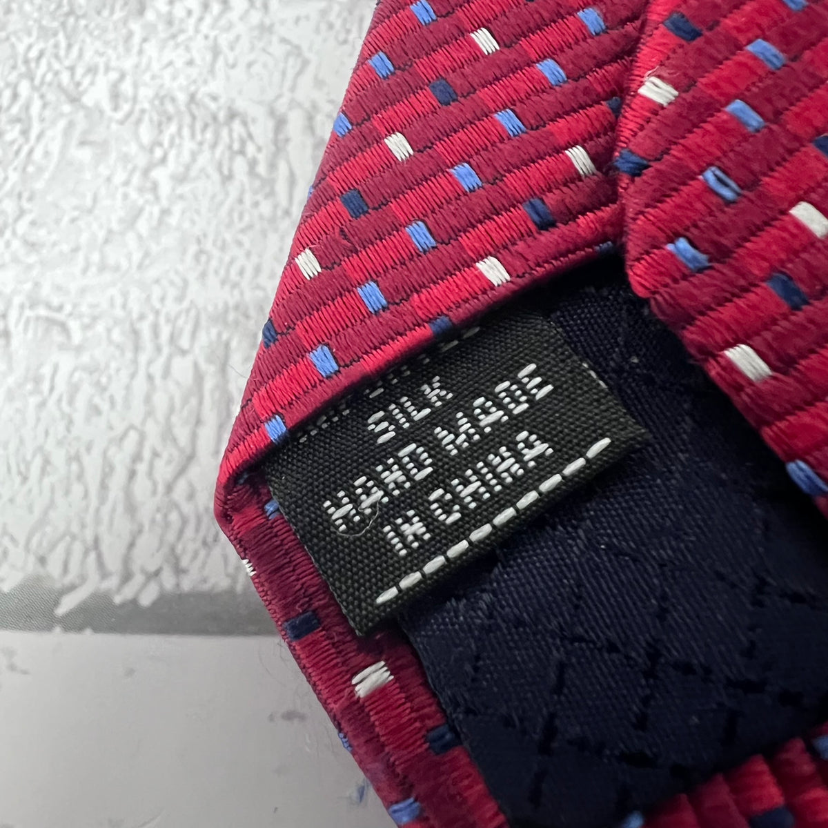 Close-up of a red silk necktie with a woven pattern of blue, white, and navy threads, showing a black fabric label that reads 'SILK HAND MADE IN CHINA'.