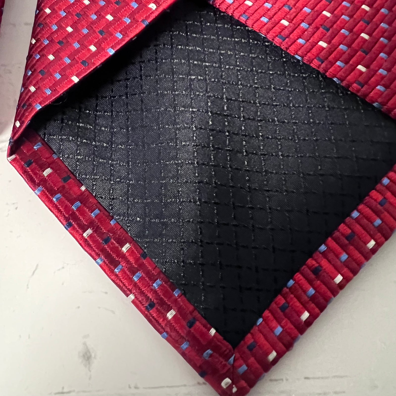 A close-up of a folded necktie showing its textured black front pattern and red woven border with small blue and white accent threads.