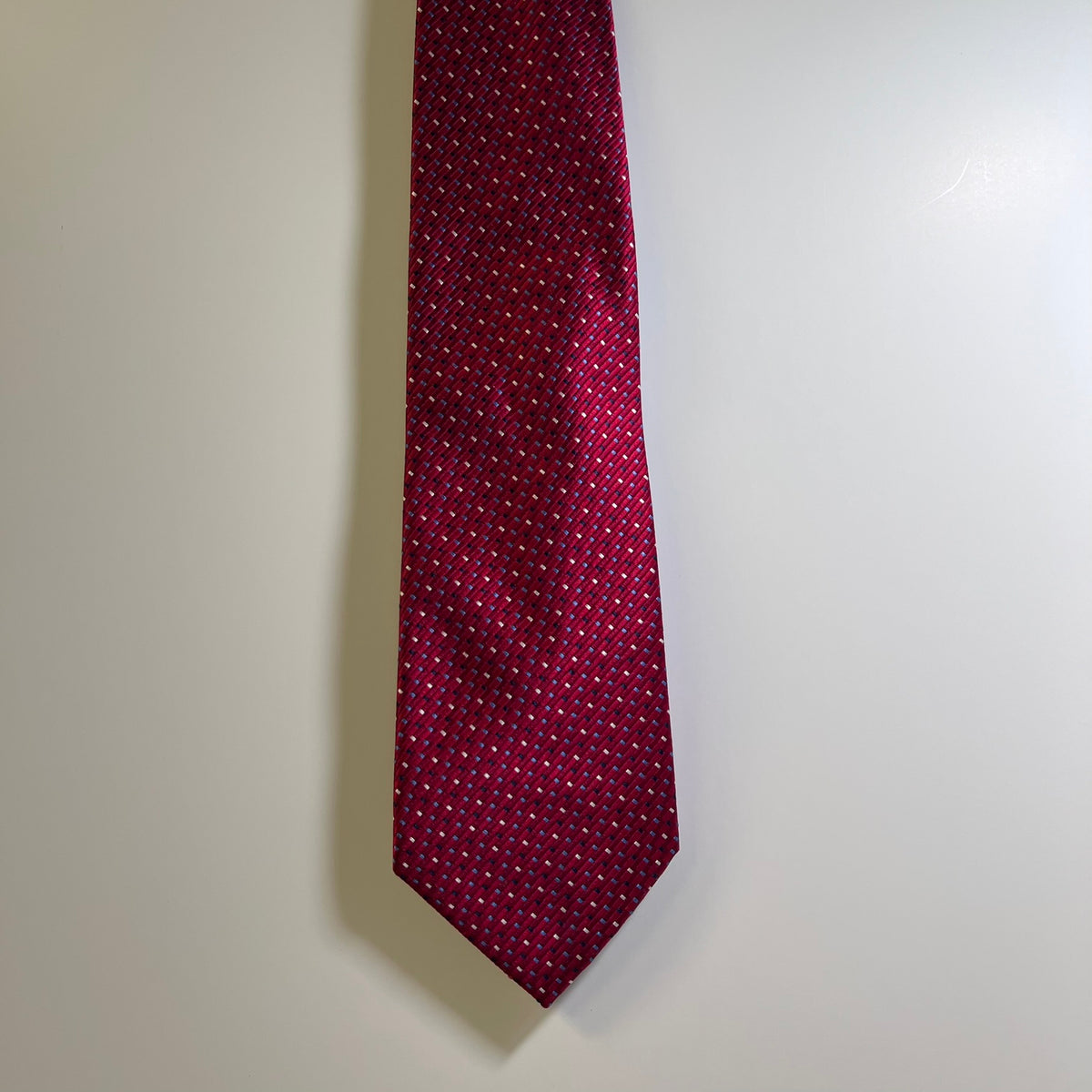 A burgundy red necktie with a small white geometric pattern, hanging against a plain off-white wall.