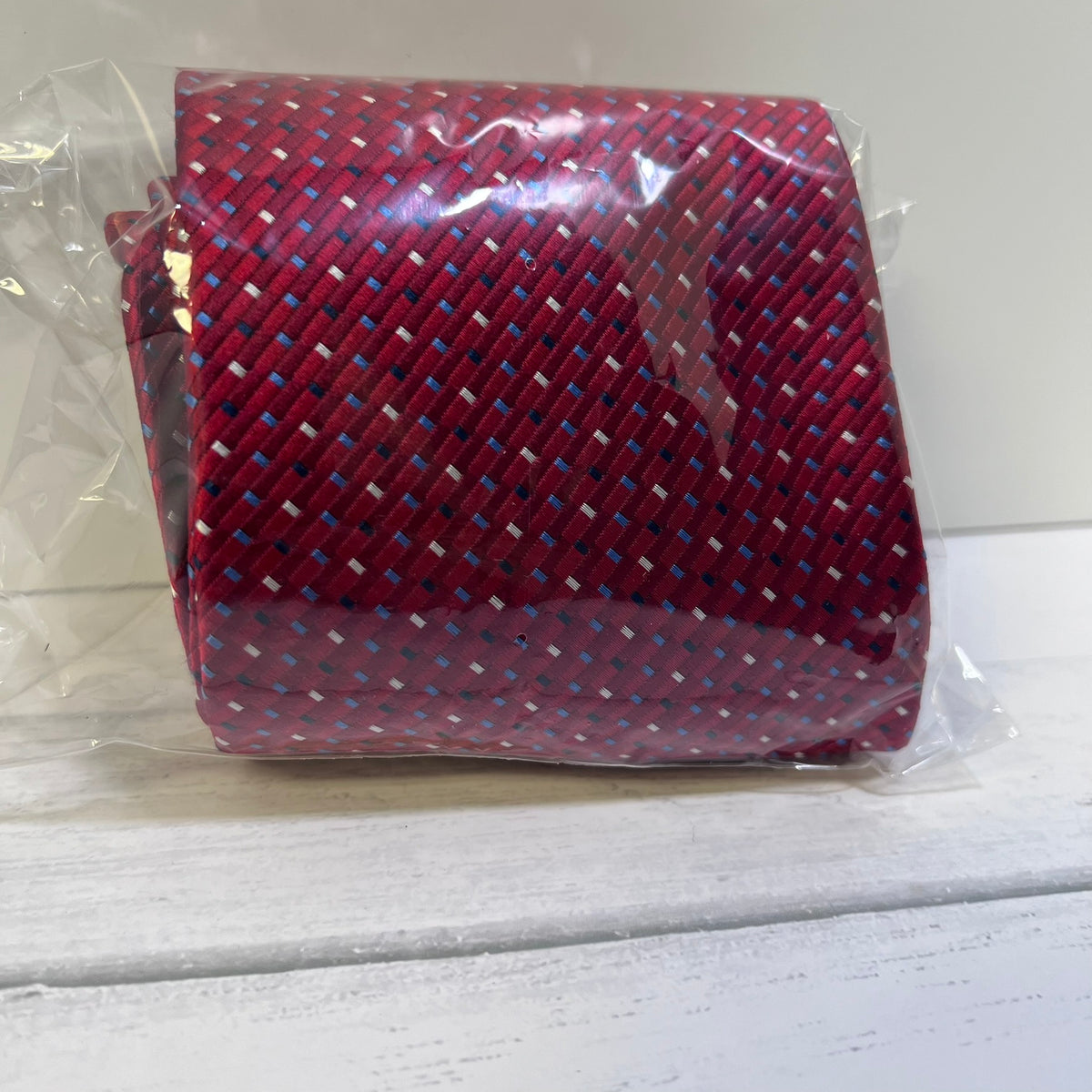 A rolled necktie in clear plastic packaging, featuring a red base with a woven pattern of blue, white, and black threads.
